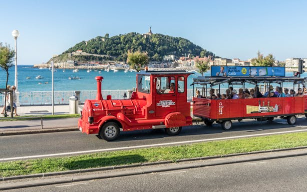Red tourist train along San Sebastian promenade with view of La Concha Bay.