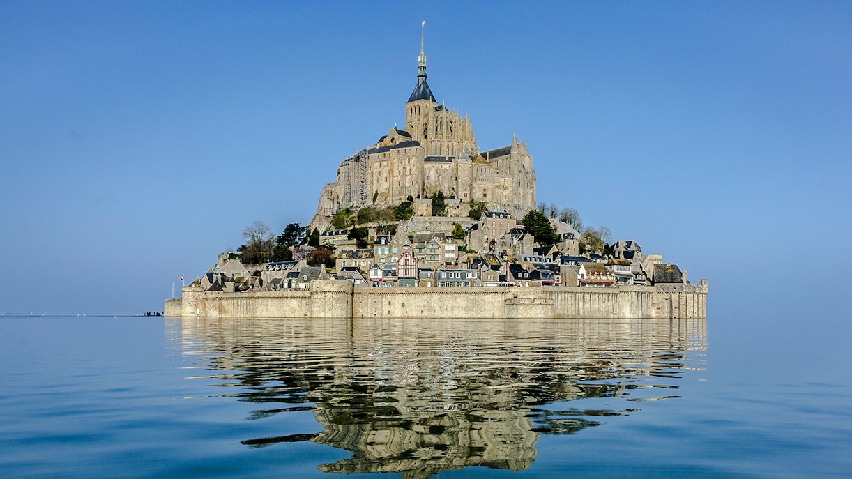 Mont Saint Michel reflected in calm water under a clear blue sky.