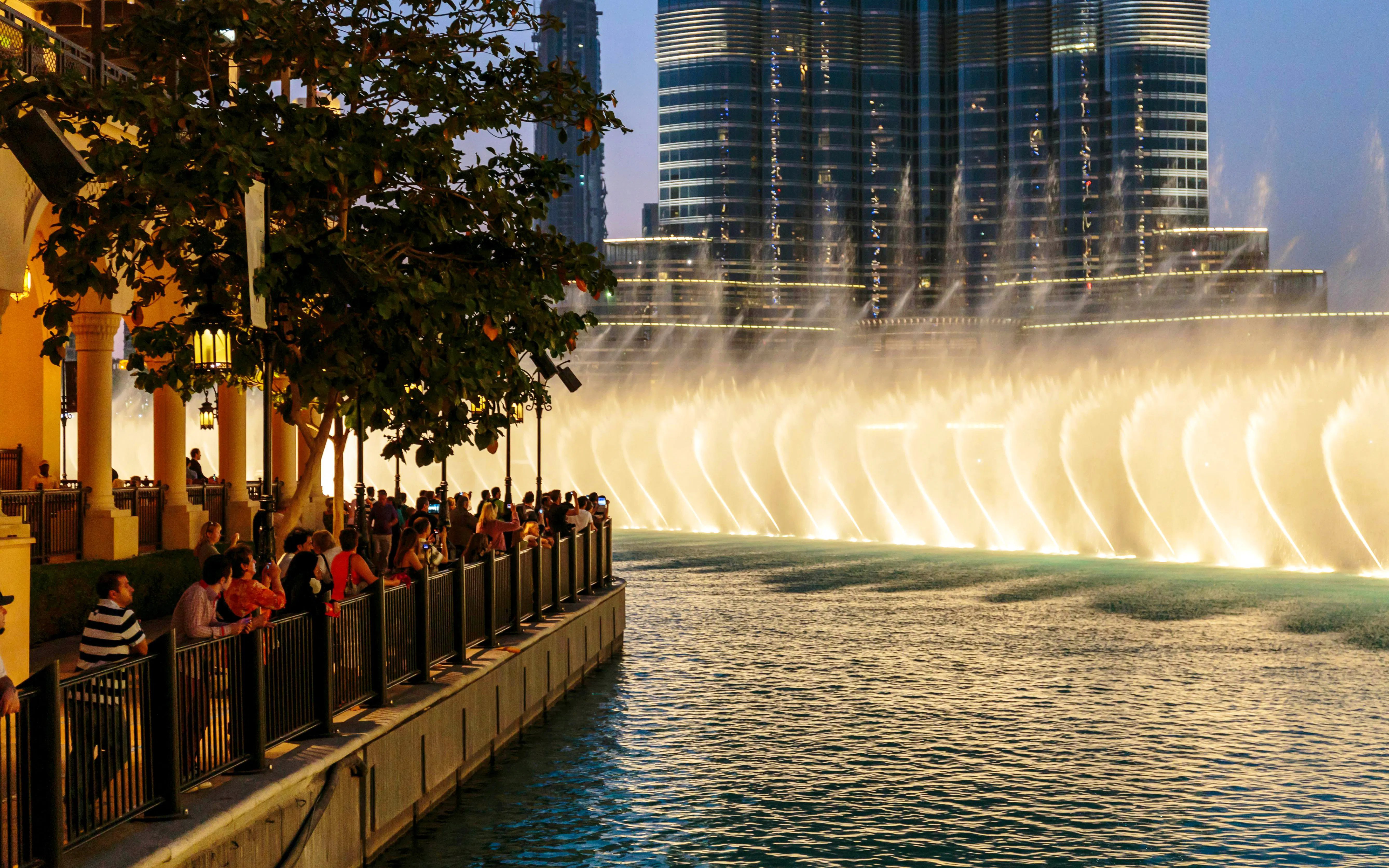 Crowd watching The Dubai Fountain show from the boardwalk with city skyline in the background.