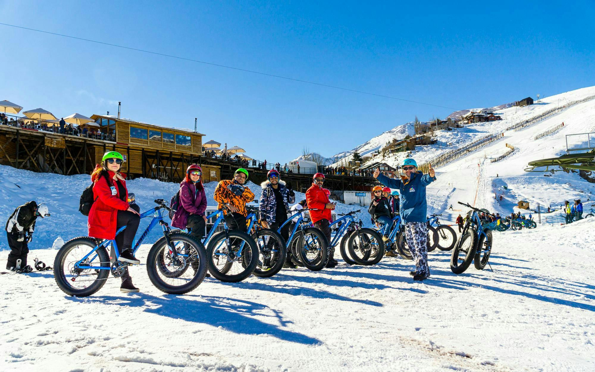 Group snow biking at Farellones Park with snowy mountains and wooden lodge in background.