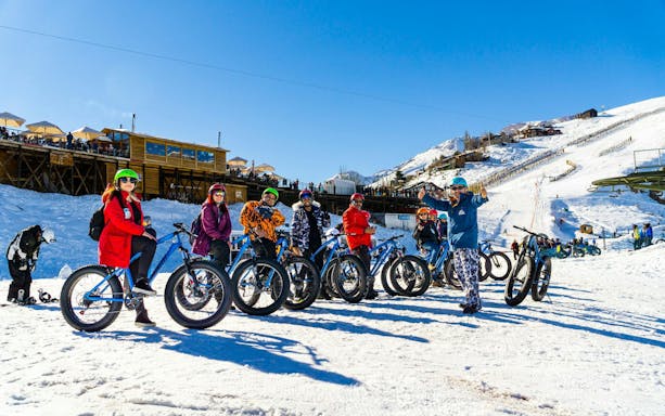 Group snow biking at Farellones Park with snowy mountains and wooden lodge in background.