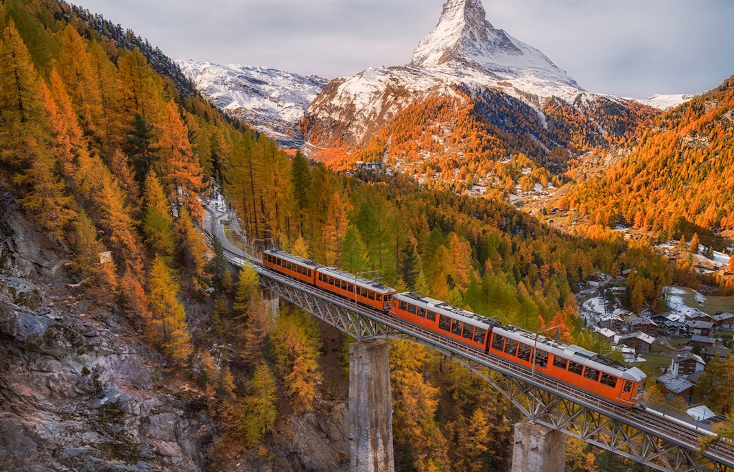 Gornergrat train crossing bridge with snowy Matterhorn peak and autumn foliage under blue cloudy sky in Zermatt, Switzerland.