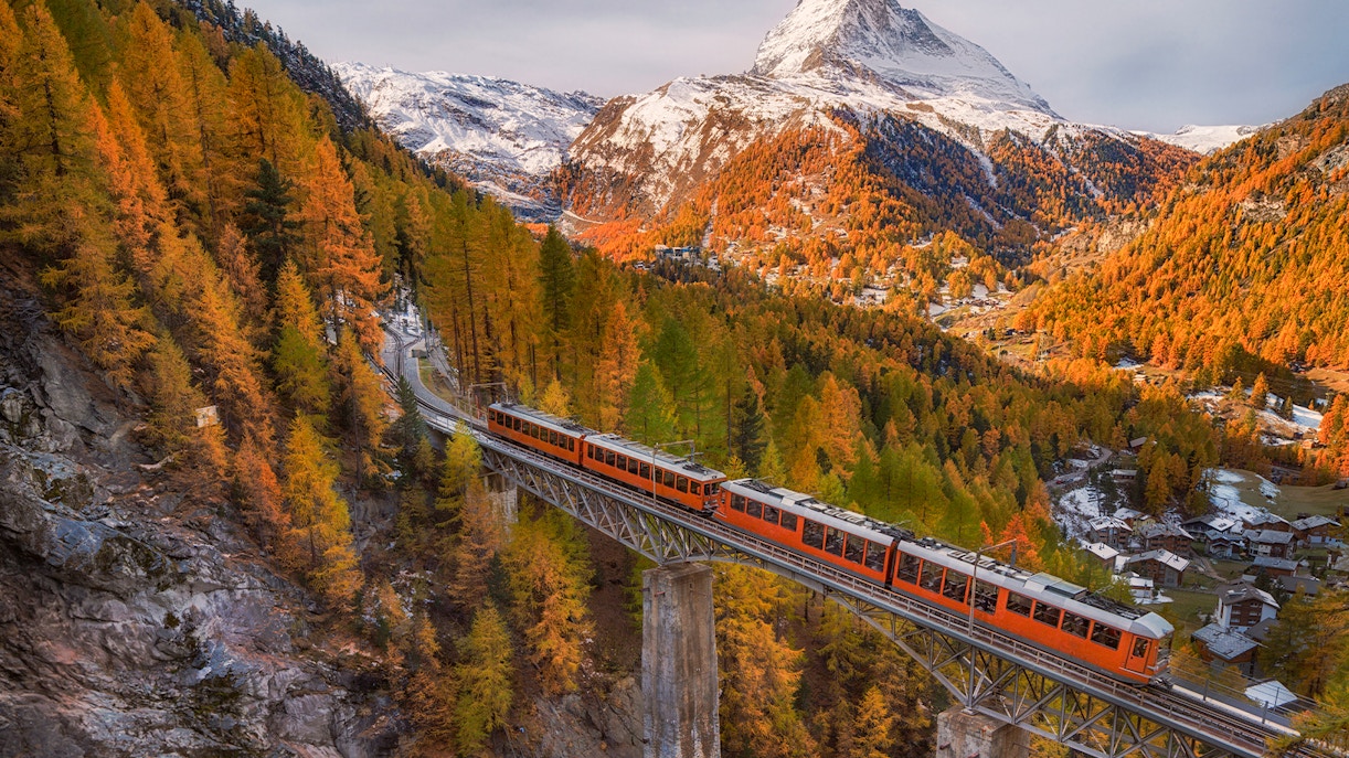 Gornergrat train crossing bridge with snowy Matterhorn peak and autumn foliage under blue cloudy sky in Zermatt, Switzerland.