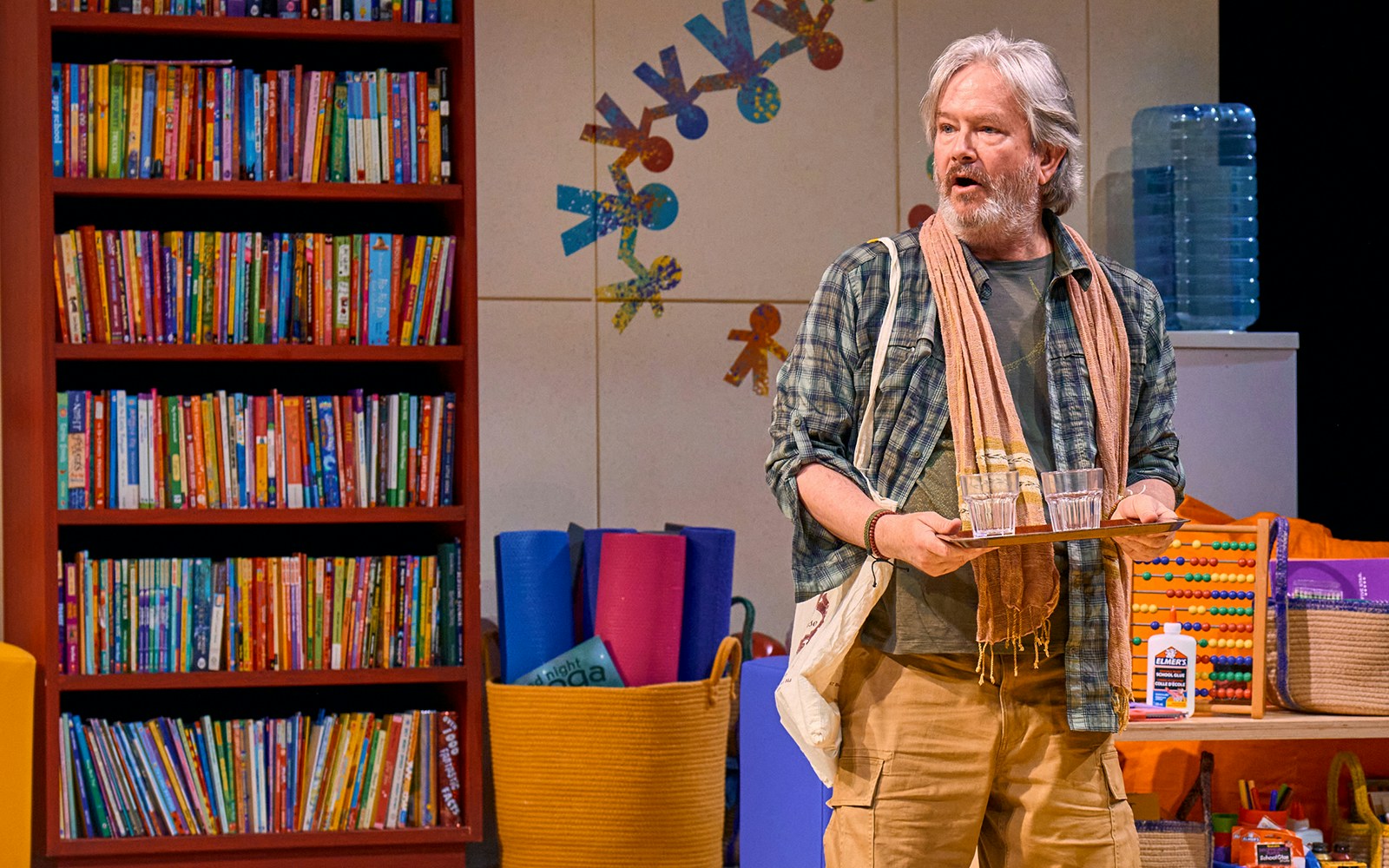 Man holding a tray with glasses in a colorful classroom setting, Eureka Day play.