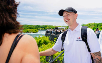 Tour guide interacting with visitors at Niagara Falls, with river and greenery in the background.