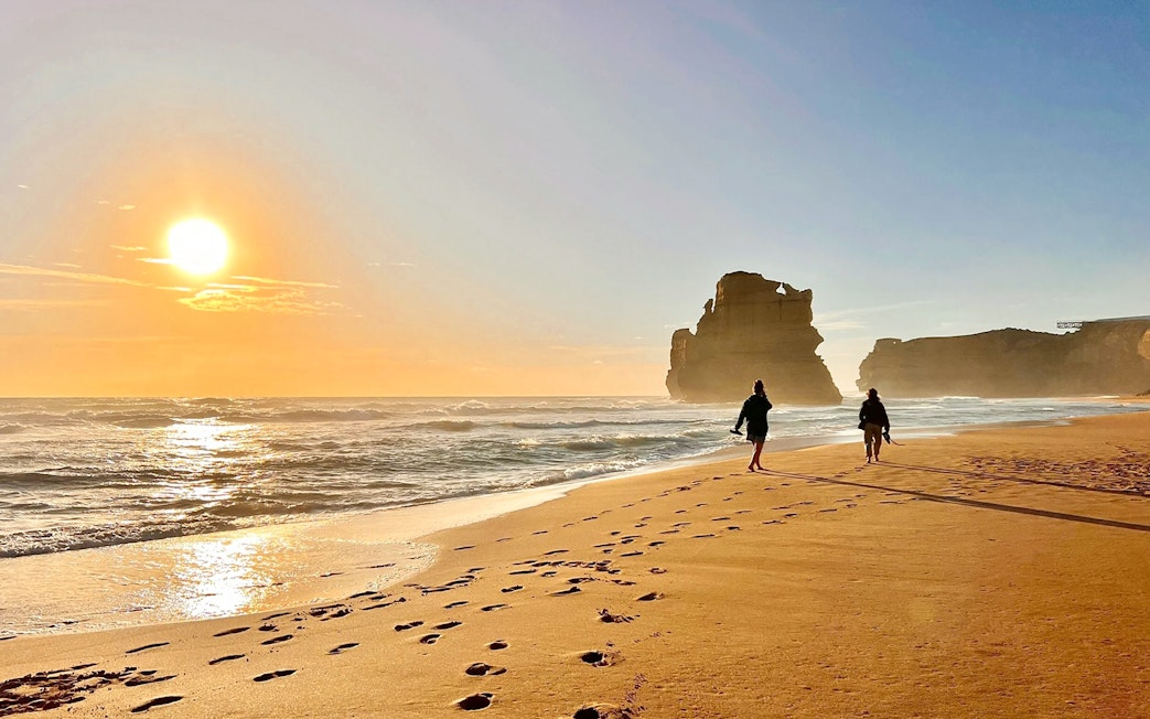 People walking on a beach at sunset near rock formations on the Great Ocean Road.