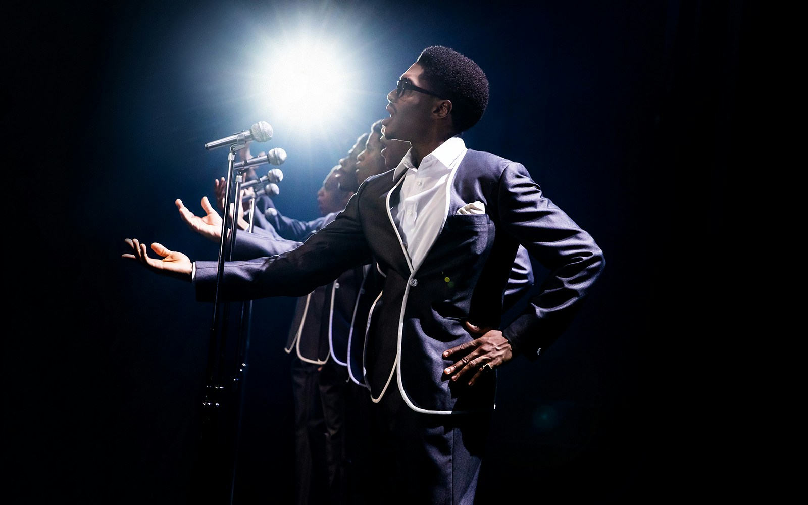 Performers in suits singing into microphones on stage during "Ain't Too Proud" show.