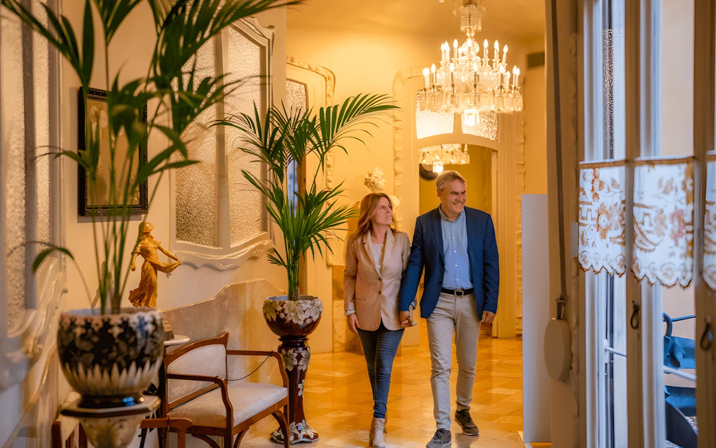 Couple walking through elegant hallway in La Padera, featuring chandeliers and decorative plants.