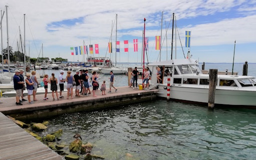 Tourists boarding a boat for a 4-hour cruise on Lake Garda to Sirmione, with flags and yachts in the background.