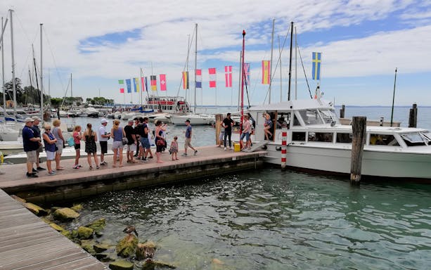 Tourists boarding a boat for a 4-hour cruise on Lake Garda to Sirmione, with flags and yachts in the background.