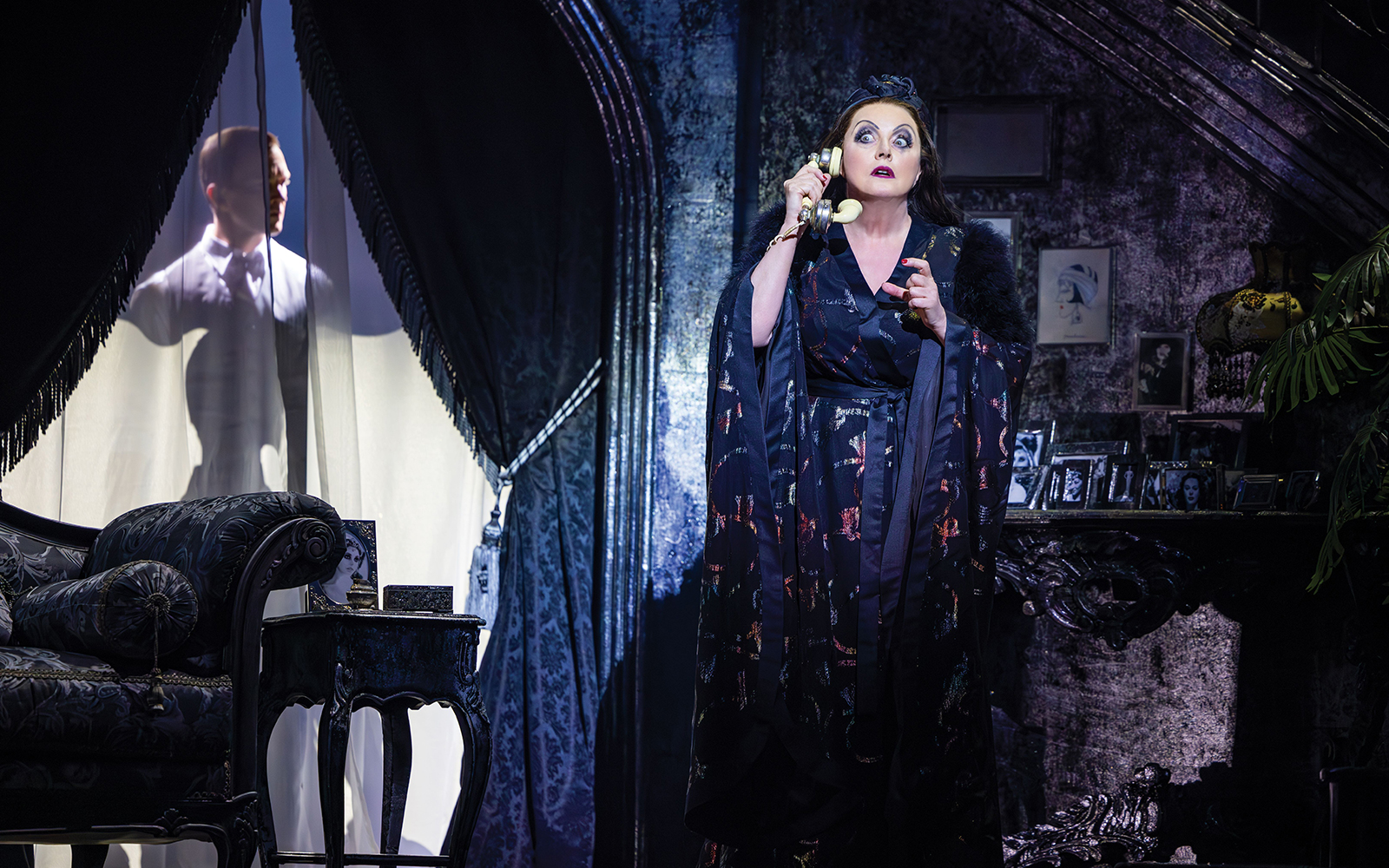 Woman in a dark room holding a phone, with a man silhouetted behind a curtain on Sunset Boulevard.