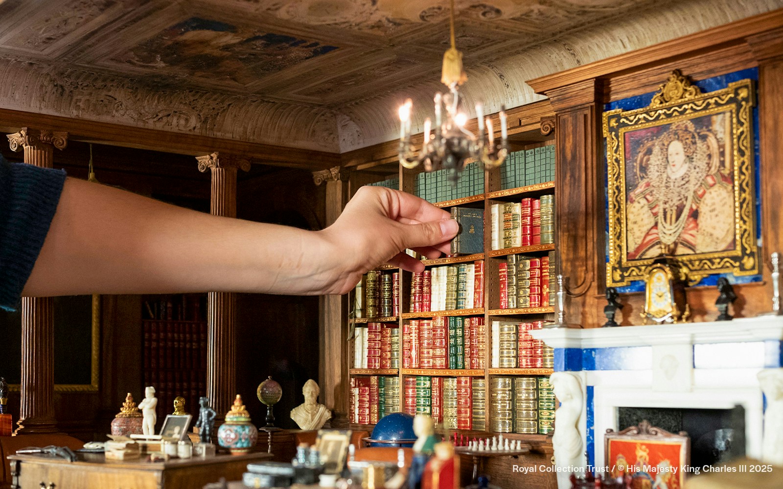 Hand arranging books in Queen Mary's Dollhouse library, featuring intricate miniature details.