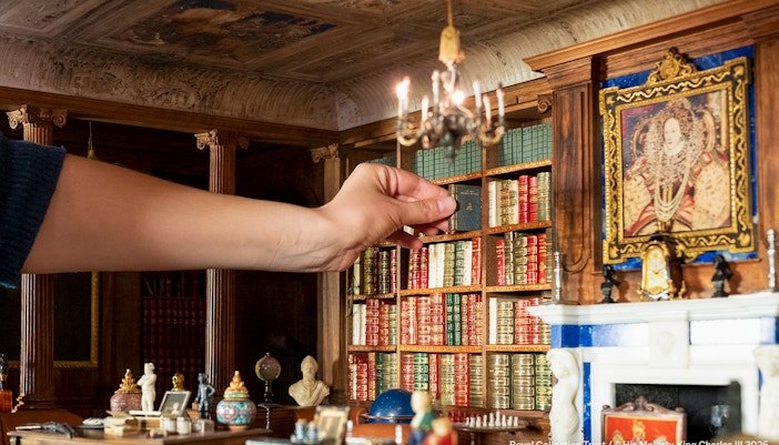 Hand arranging books in Queen Mary's Dollhouse library, featuring intricate miniature details.