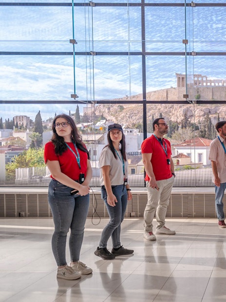 Visitors on a guided tour in Acropolis Museum with view of the Parthenon through large windows.