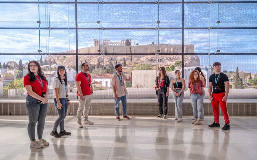 Visitors on a guided tour in Acropolis Museum with view of the Parthenon through large windows.