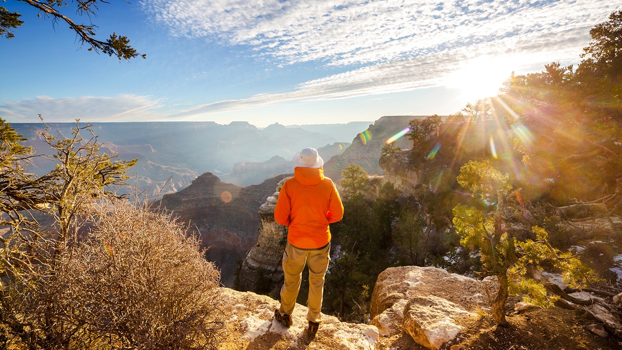 Person standing on Grand Canyon edge at sunrise, overlooking vast canyon landscape.