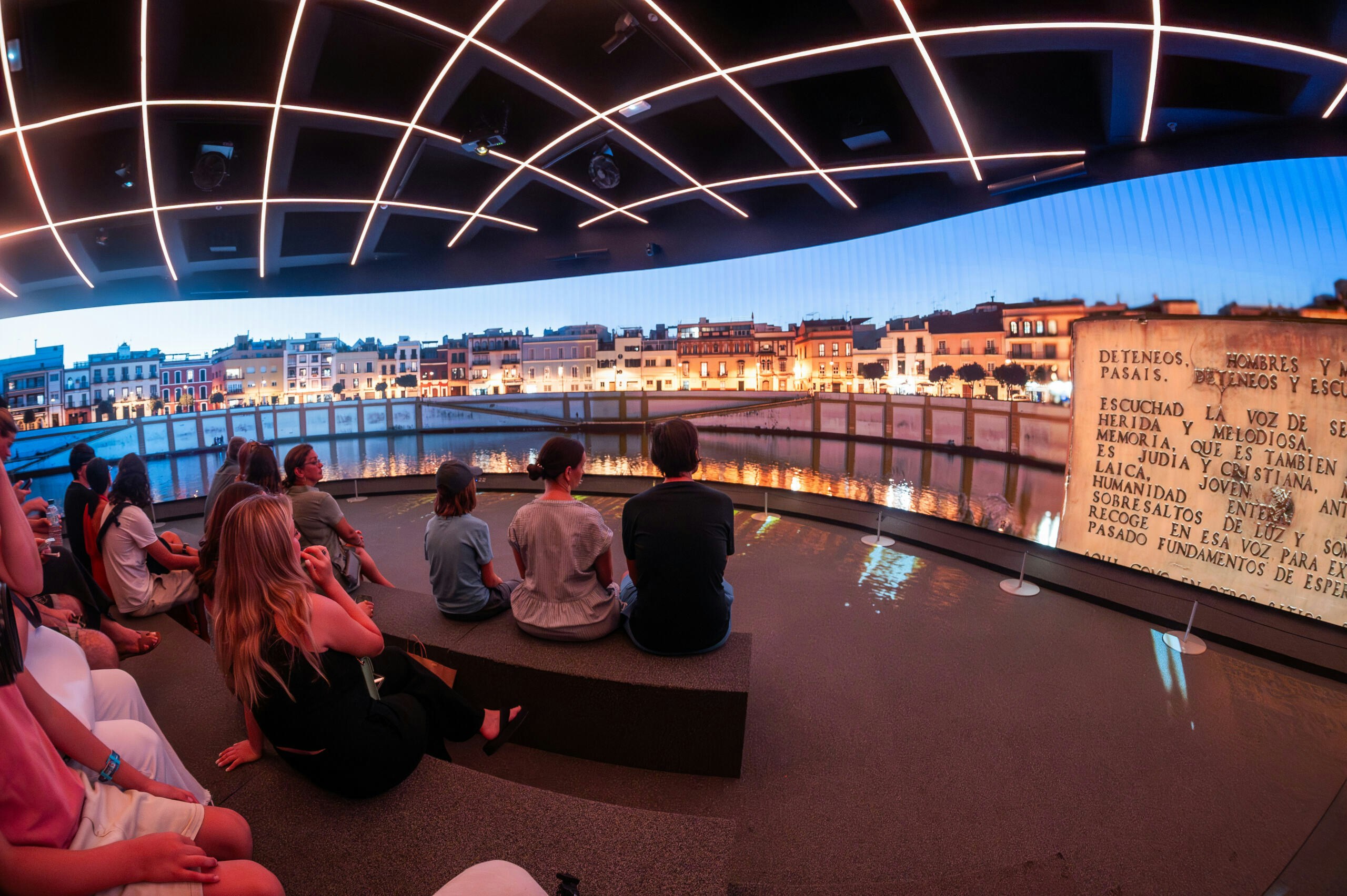 Visitors seated inside Setas de Sevilla, Spain, watching a night-time cityscape display.