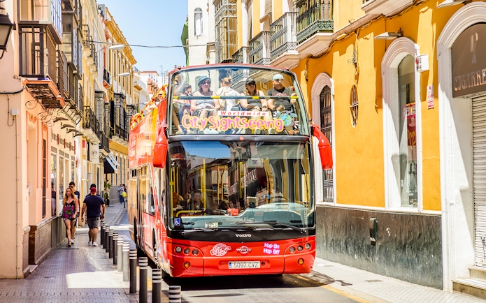 Seville hop-on hop-off bus on a narrow street with tourists on board.