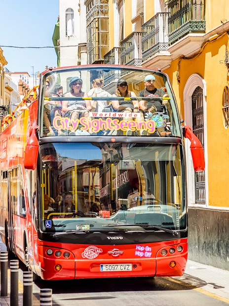 Seville hop-on hop-off bus on a narrow street with tourists on board.