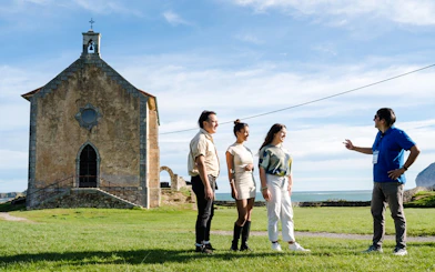 Tour group at chapel near Dragonstone, Mundaka, with ocean view in background.