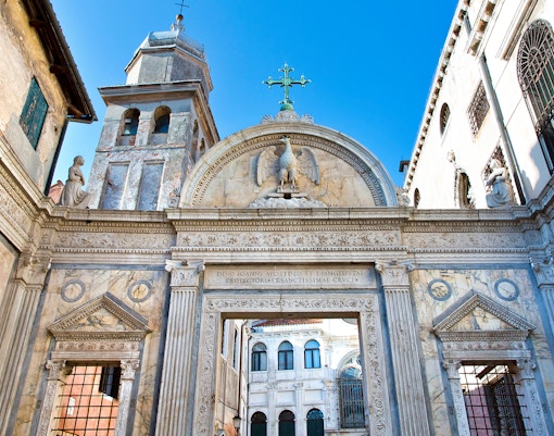 White stone gate with ornaments and weathered medieval stone facades of Scuola Grande di San Giovanni Evangelista at City of Venice on a cloudy summer day