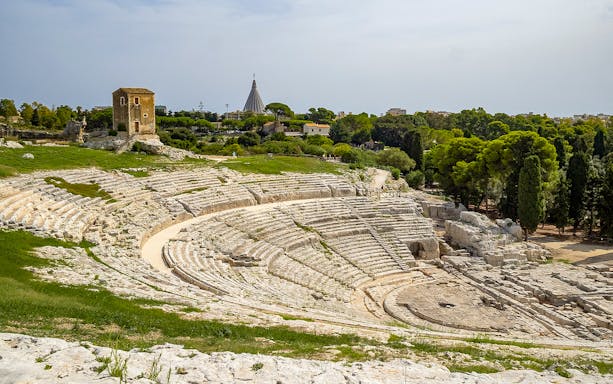 Ancient Greek theater in Neapolis Archaeological Park, Syracuse, with surrounding greenery.