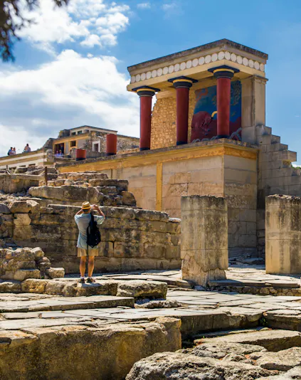 Knossos Palace ruins with red columns on Crete, Greece, visitor exploring site.