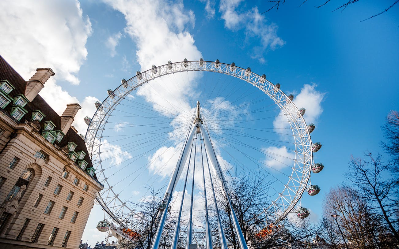 London Eye against a blue sky with nearby building and trees.