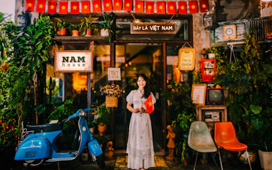 Woman holding Vietnamese flag outside Nam House café in Da Nang, with motorbike and plants.