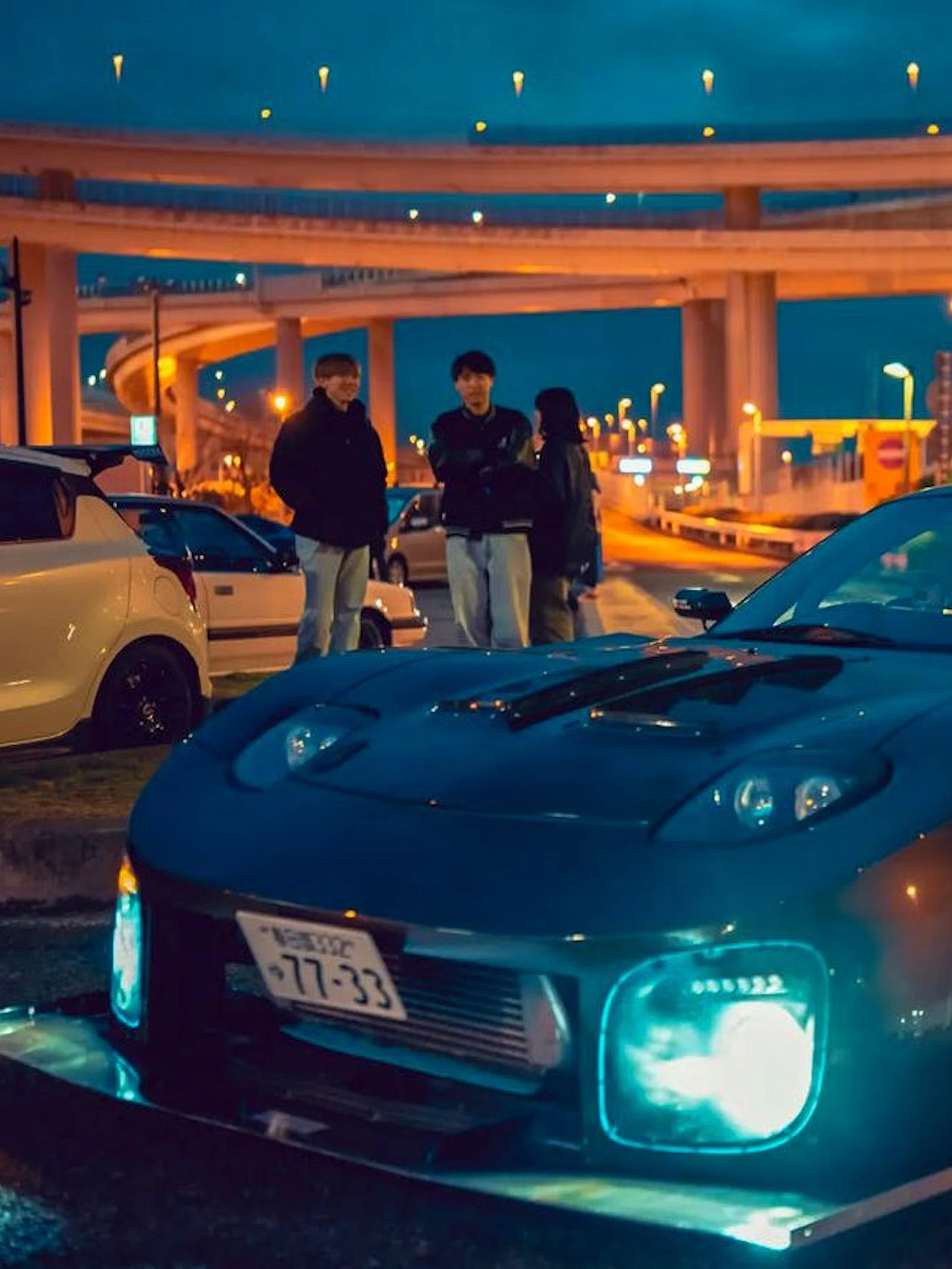 Car with headlights on at Daikoku PA Car Meeting, Tokyo, with illuminated highway in background.