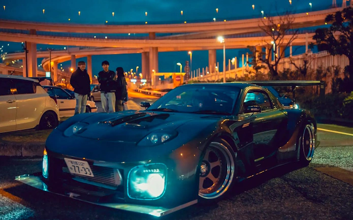 Car with headlights on at Daikoku PA Car Meeting, Tokyo, with illuminated highway in background.