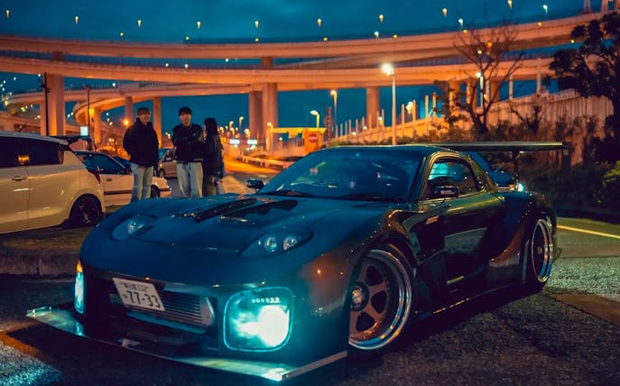 Car with headlights on at Daikoku PA Car Meeting, Tokyo, with illuminated highway in background.