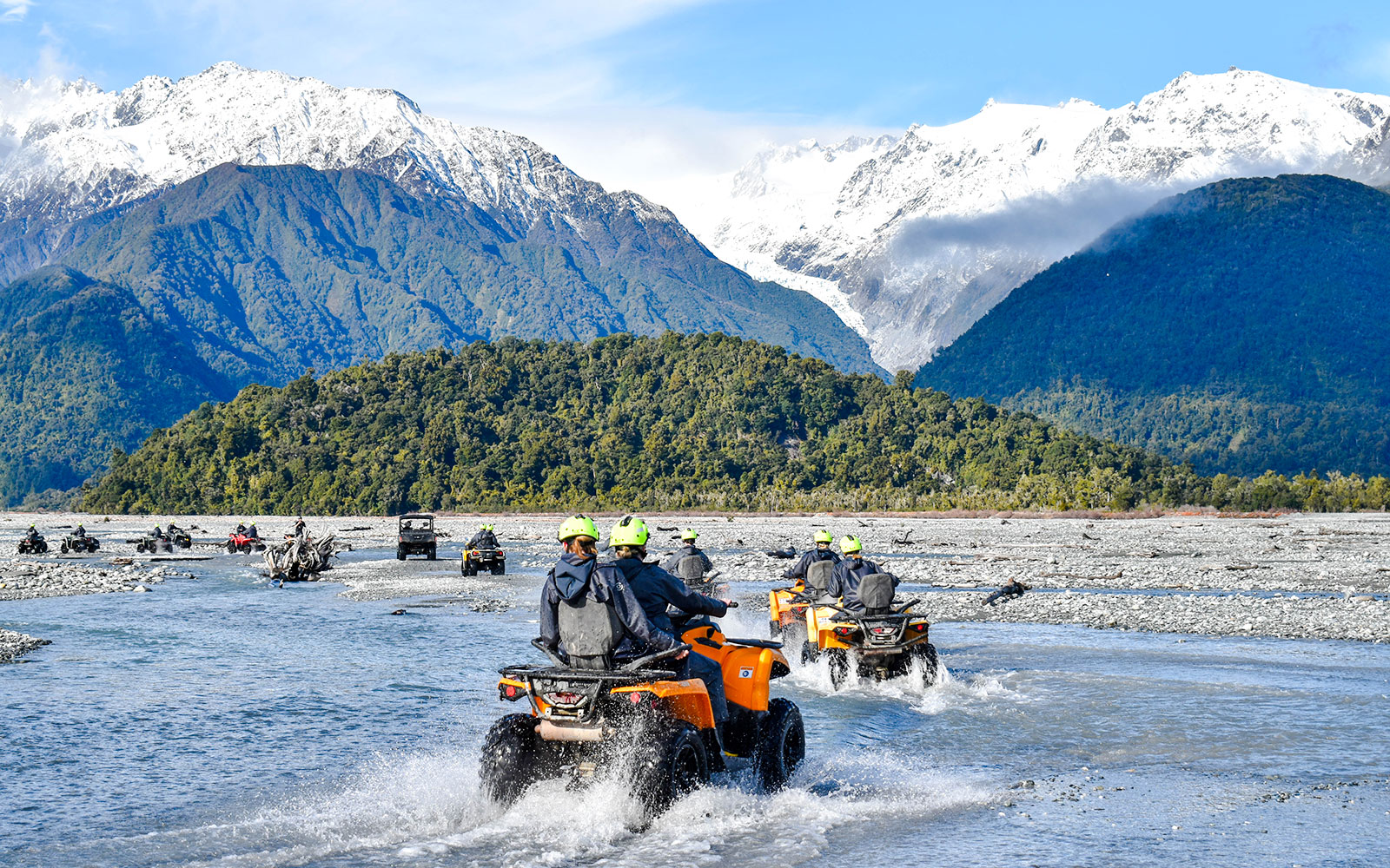 Quad biking through river with snow-capped mountains at Franz Josef, New Zealand.