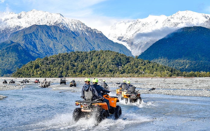 Quad biking through river with snow-capped mountains at Franz Josef, New Zealand.