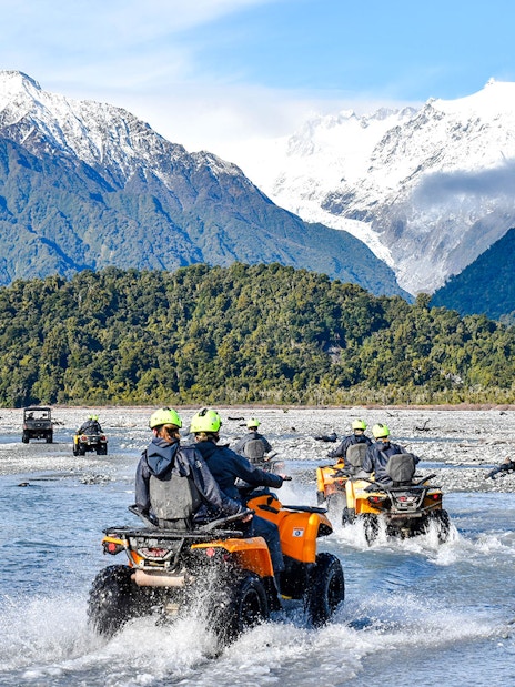 Quad biking through river with snow-capped mountains at Franz Josef, New Zealand.