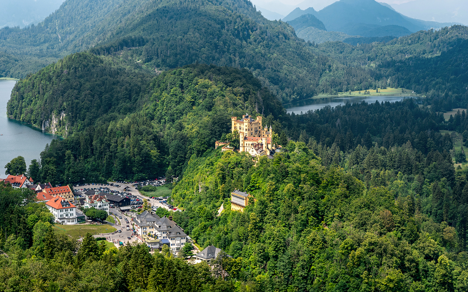 Hohenschwangau Castle surrounded by lush forest and mountains, view of nearby village and lake.