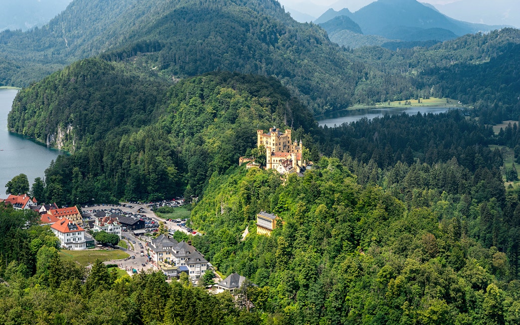 Hohenschwangau Castle surrounded by lush forest and mountains, view of nearby village and lake.
