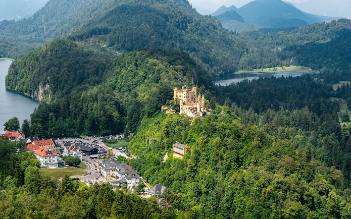 Hohenschwangau Castle surrounded by lush forest and mountains, view of nearby village and lake.