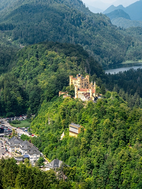 Hohenschwangau Castle surrounded by lush forest and mountains, view of nearby village and lake.