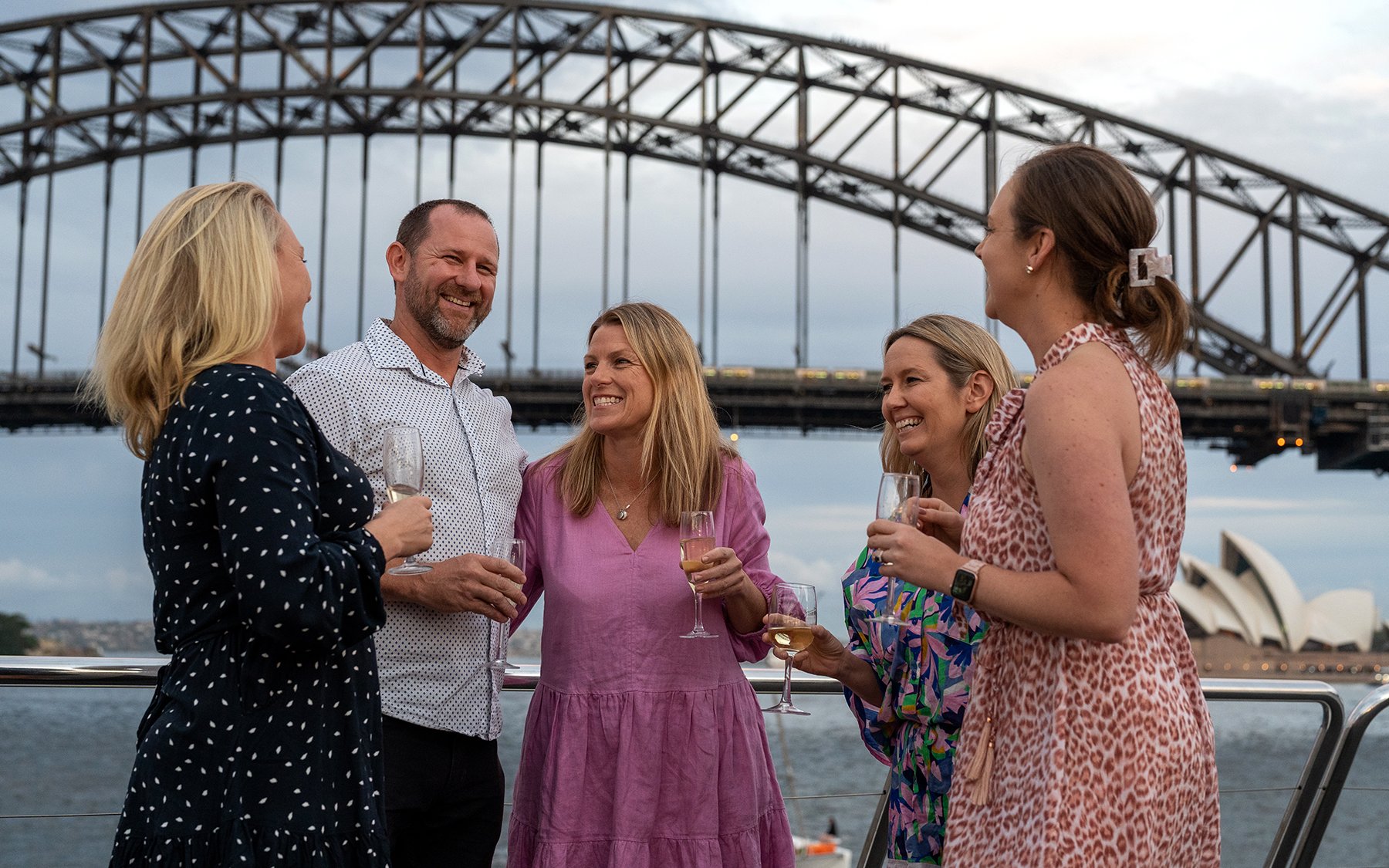 Group enjoying wine on Sydney Harbour cruise with Harbour Bridge and Opera House in background.