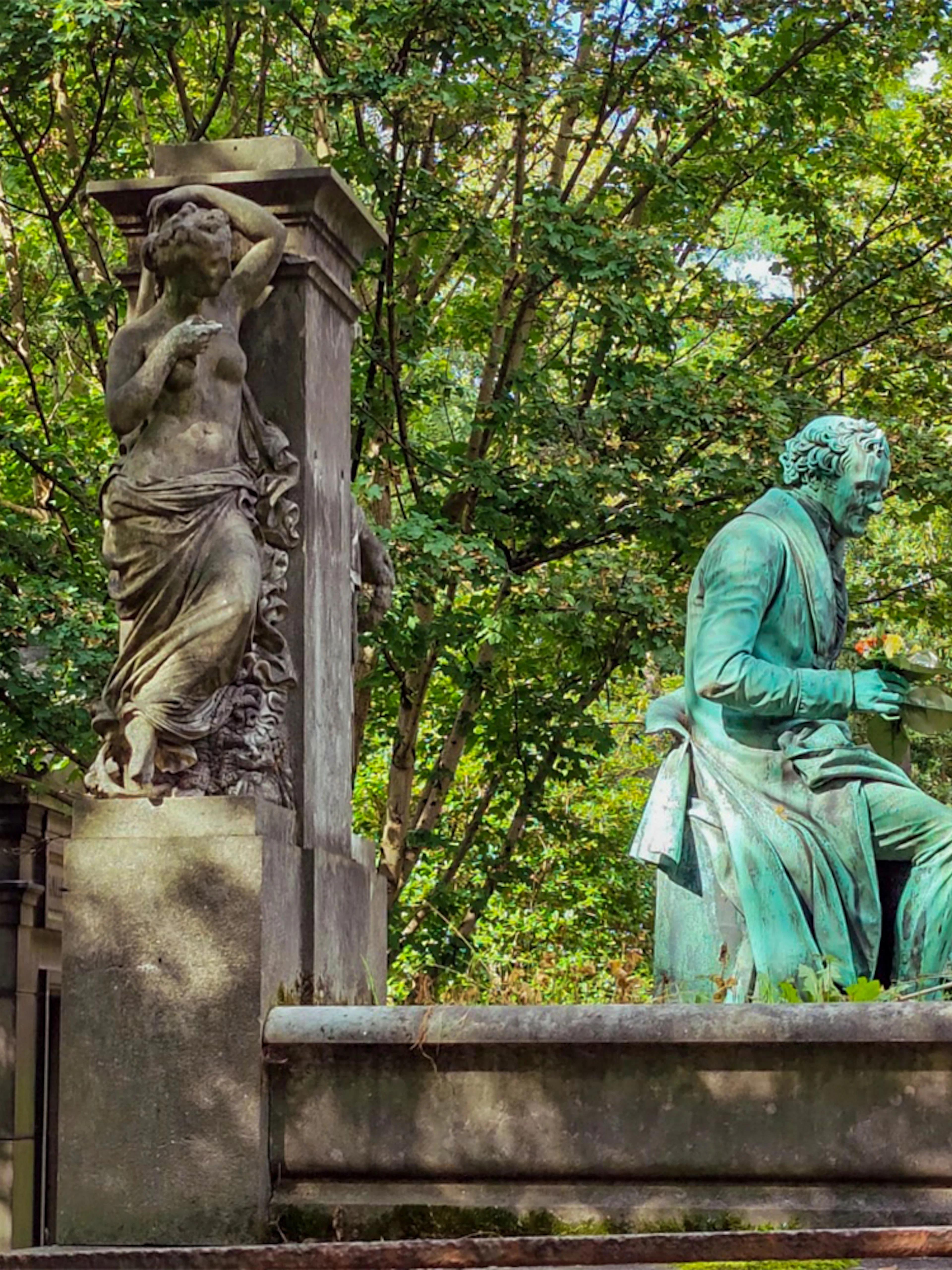 Statue of a seated figure and a standing sculpture at Père Lachaise Cemetery, Paris.