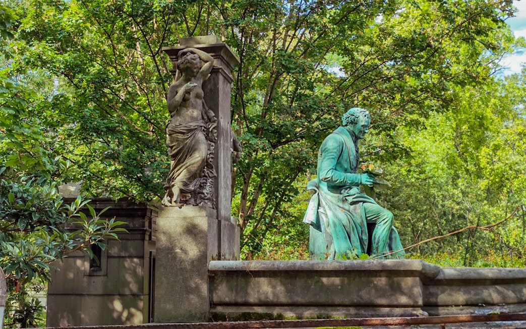 Statue of a seated figure and a standing sculpture at Père Lachaise Cemetery, Paris.