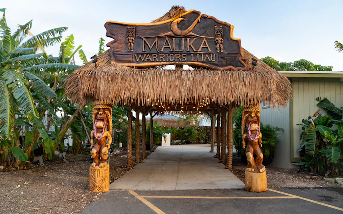 Entrance arch with dried grass at Mauka Warriors Luau, featuring carved wooden statues.