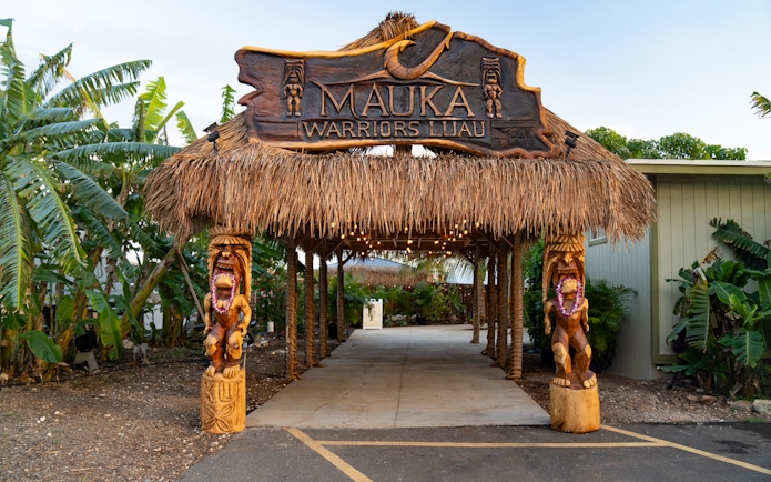 Entrance arch with dried grass at Mauka Warriors Luau, featuring carved wooden statues.
