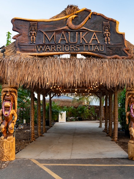 Entrance arch with dried grass at Mauka Warriors Luau, featuring carved wooden statues.