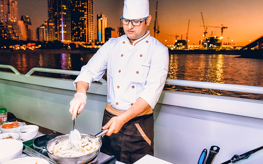 Chef preparing meal on Mega Yacht Dinner Cruise in Dubai with city skyline at sunset.
