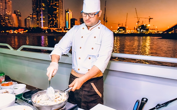Chef preparing meal on Mega Yacht Dinner Cruise in Dubai with city skyline at sunset.