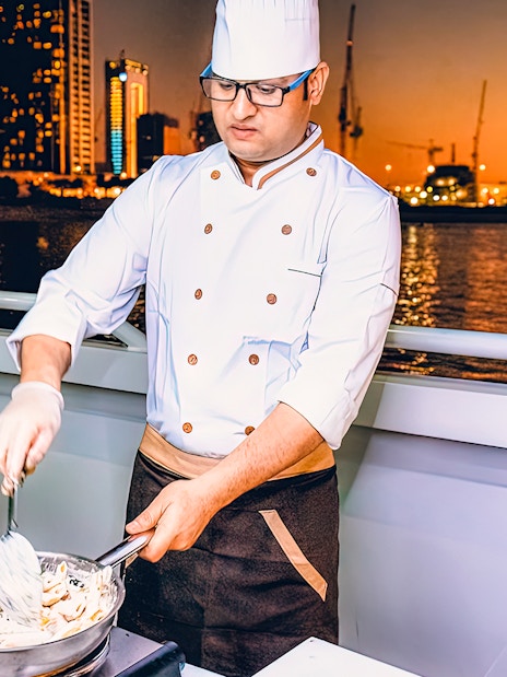 Chef preparing meal on Mega Yacht Dinner Cruise in Dubai with city skyline at sunset.