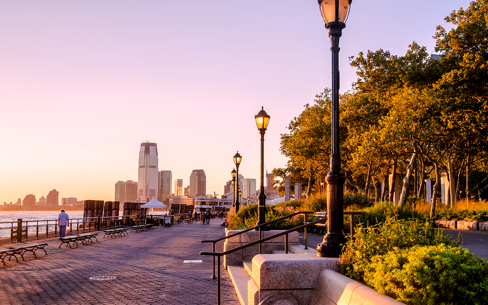 Battery Park promenade at sunset with New York City skyline in the background.