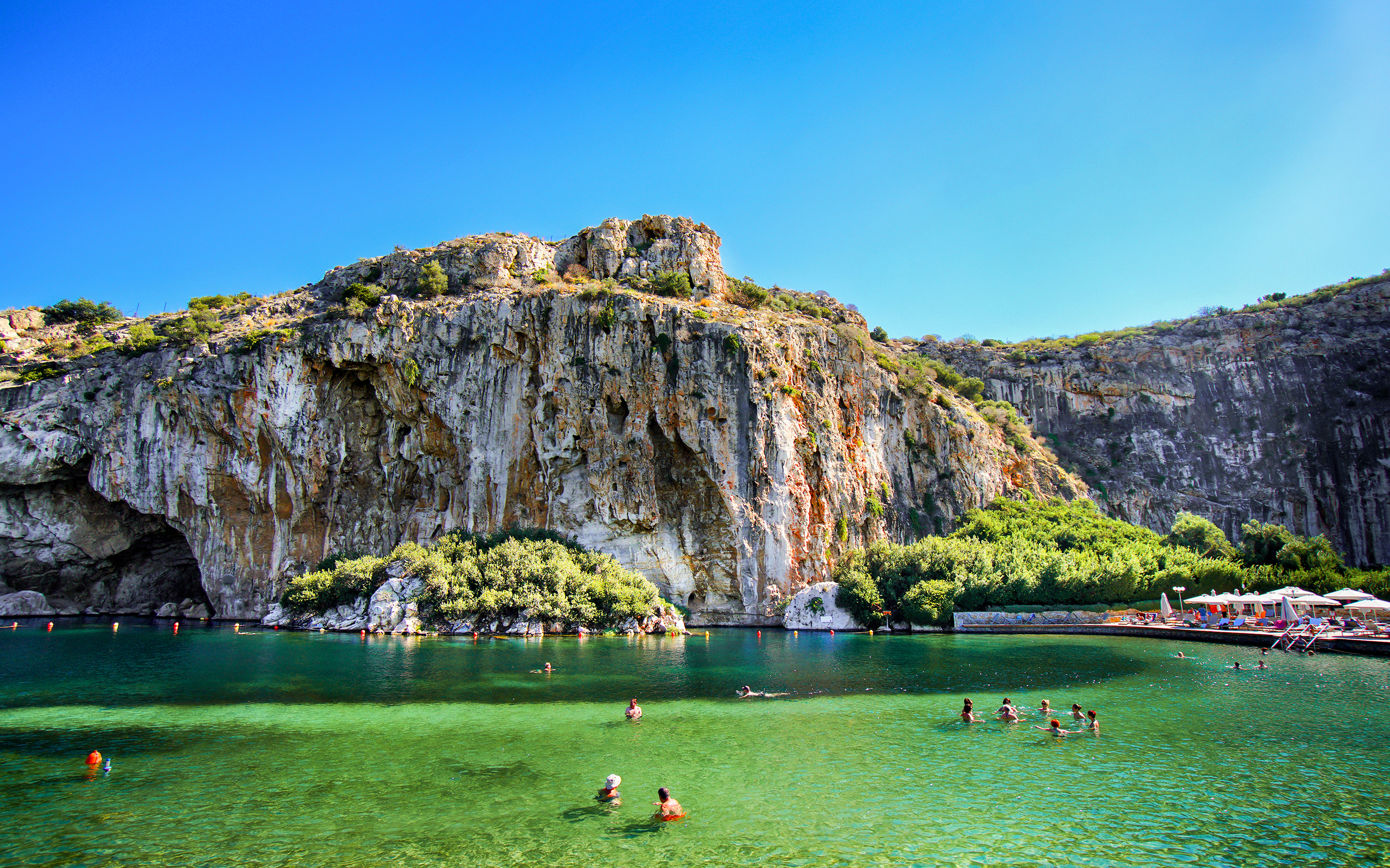 Swimmers enjoying Vouliagmeni Lake near Athens with rocky cliffs and greenery.